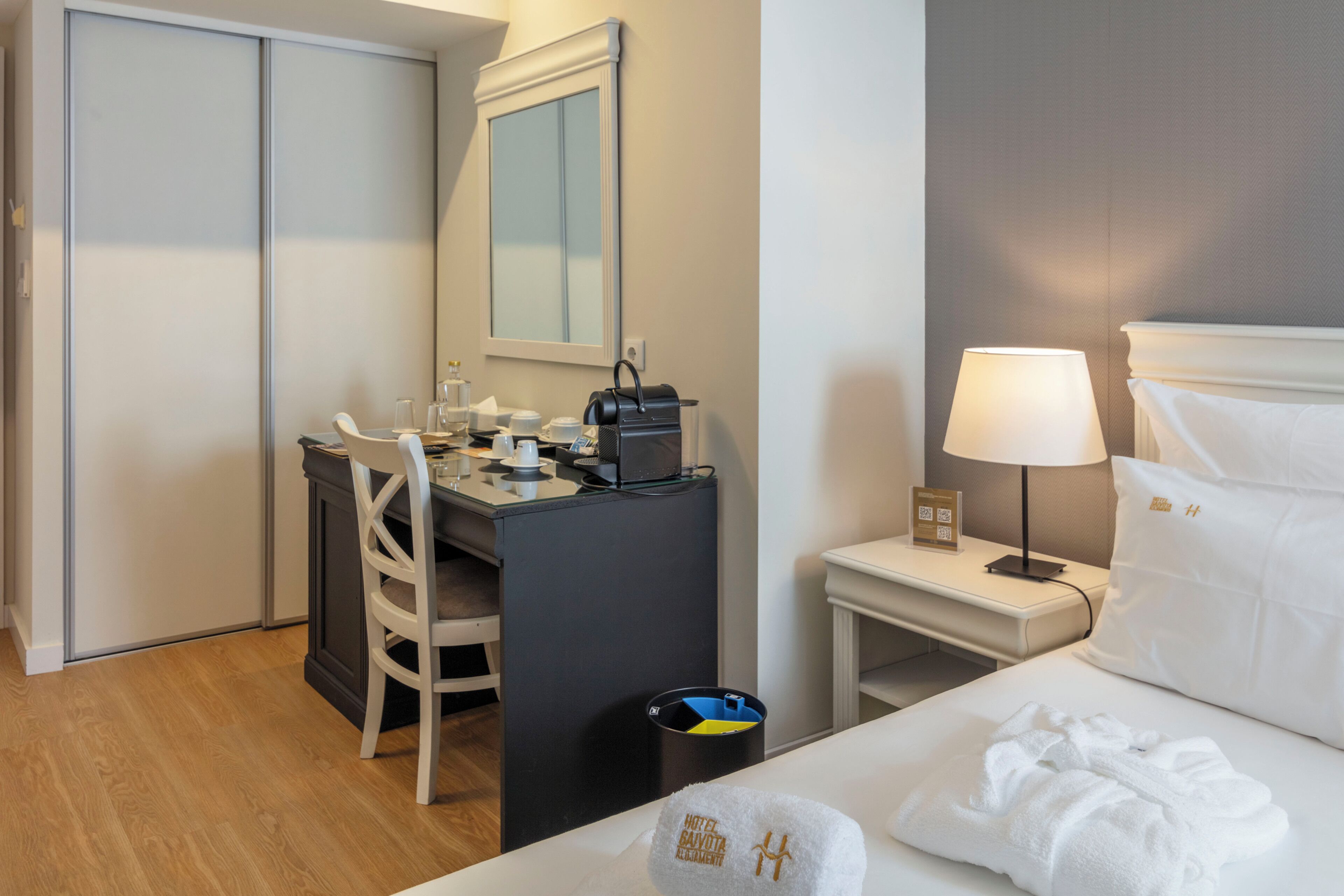 A hotel room with a bed, a desk, a chair, a lamp, and a trash bin.
