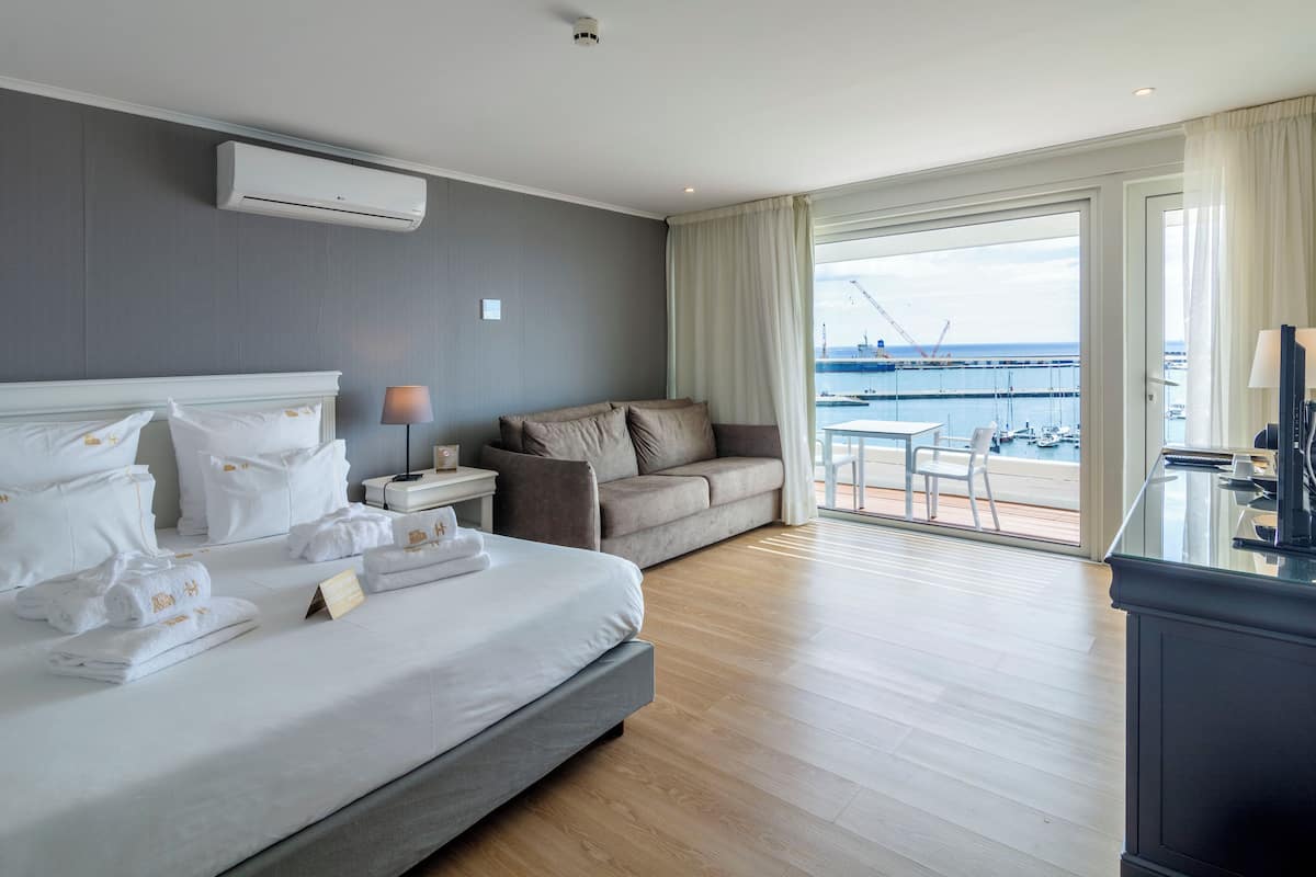 A hotel room with a large bed, a sofa, a balcony with a view of the sea, and a desk with a lamp.