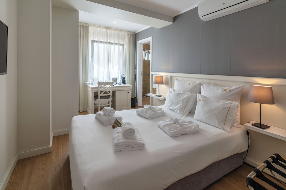 A hotel room with a neatly made bed, a desk with a chair, a mirror, and a bedside table with a lamp.