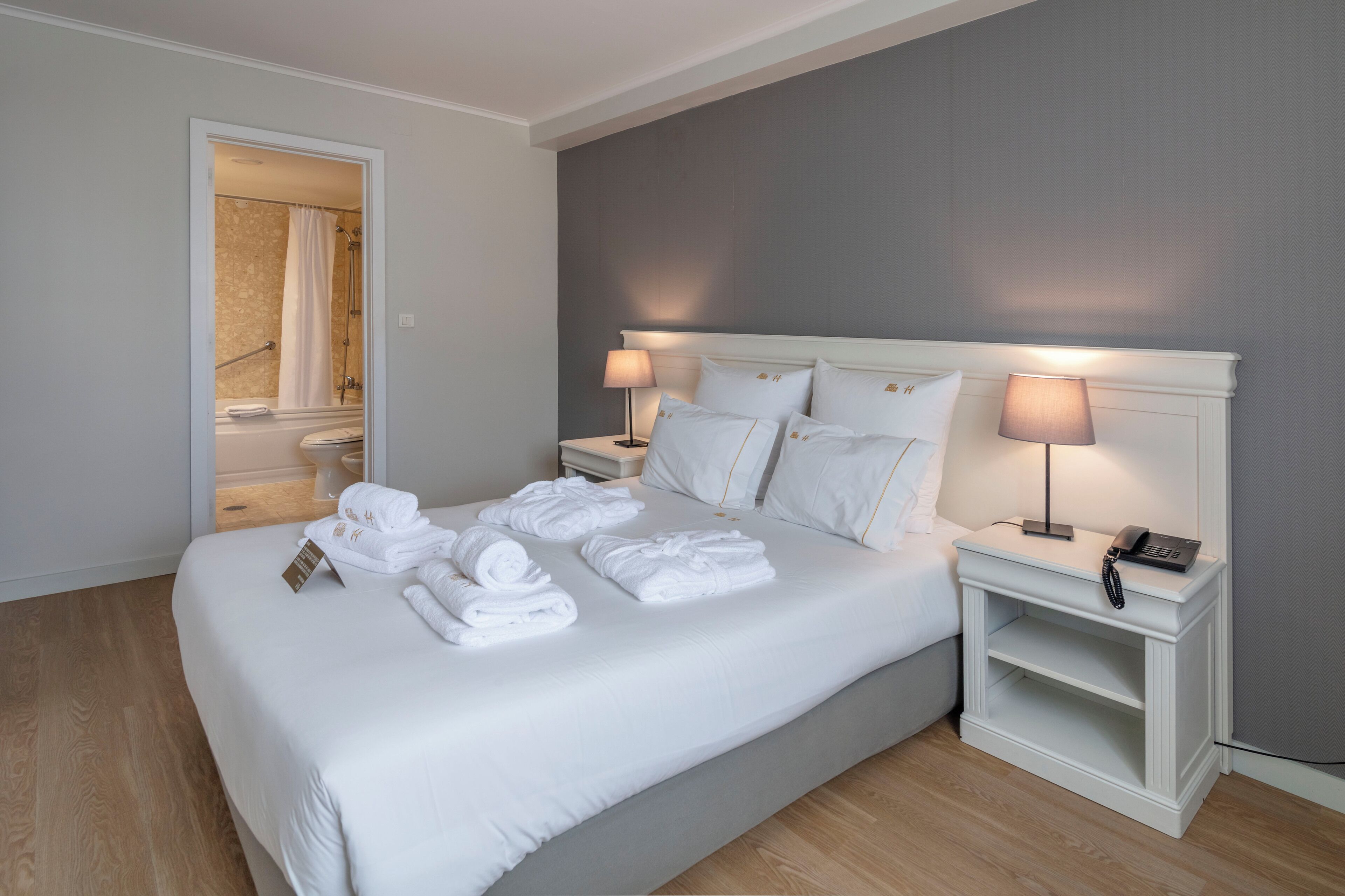 Een net opgemaakt bed met witte lakens en handdoeken, een nachtkastje met een lamp en een telefoon.