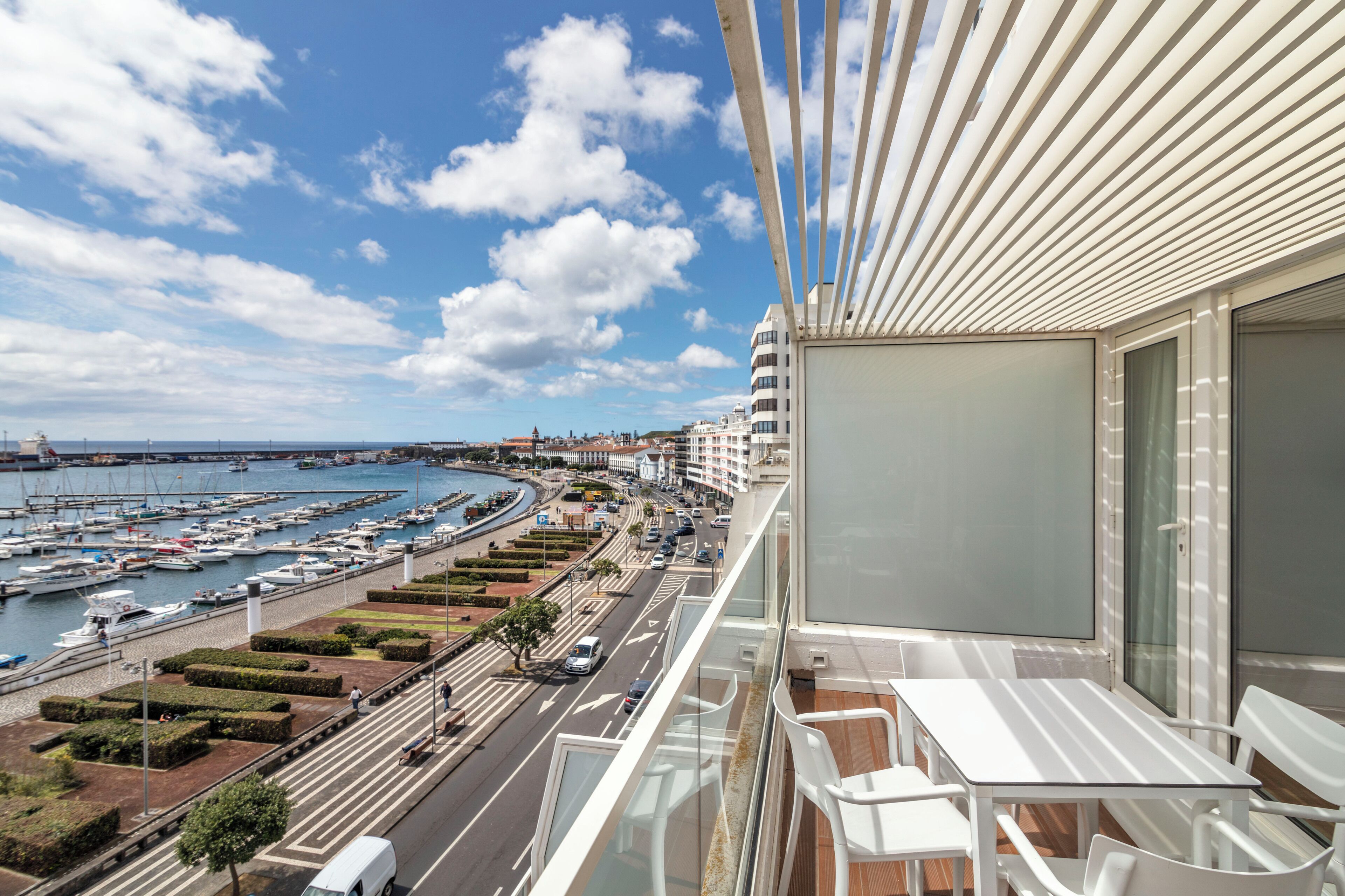 Un balcón con vista a un puerto deportivo, una carretera con vehículos y un edificio moderno con fachada de vidrio.