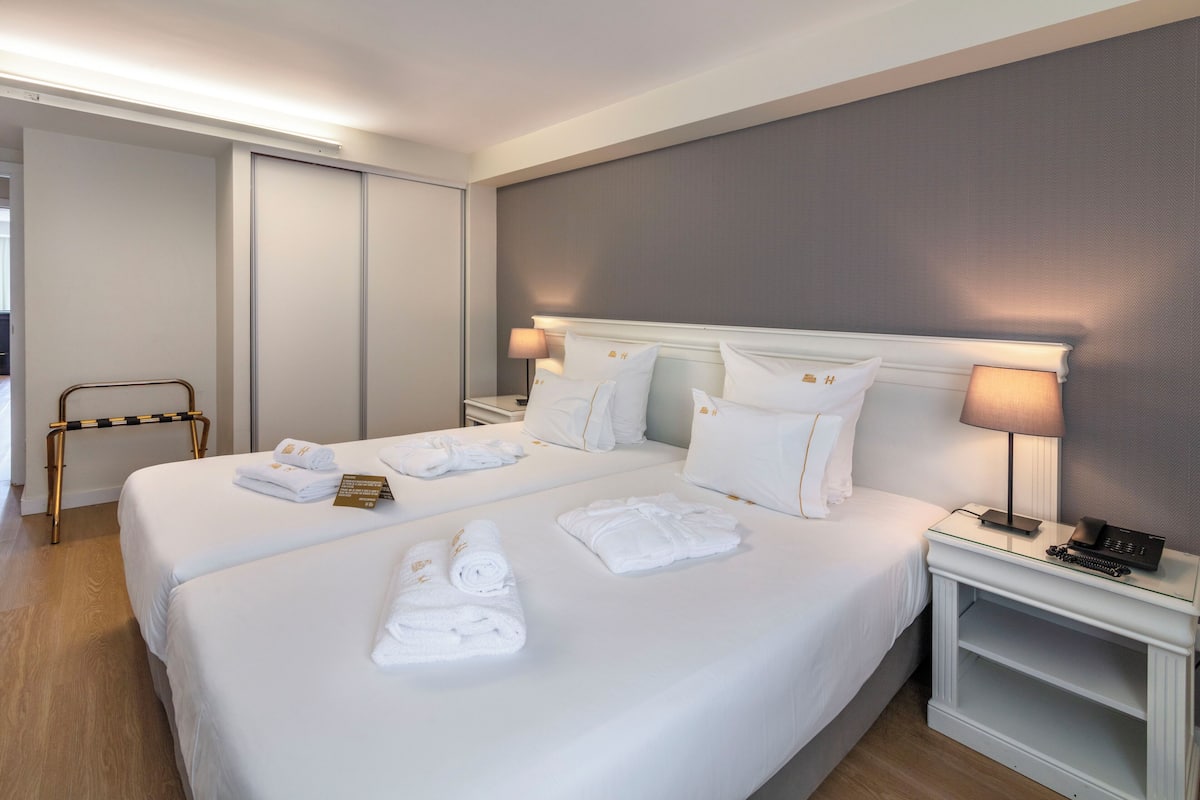 A hotel room with two beds, white linens, a bedside table with a lamp, and a chair.