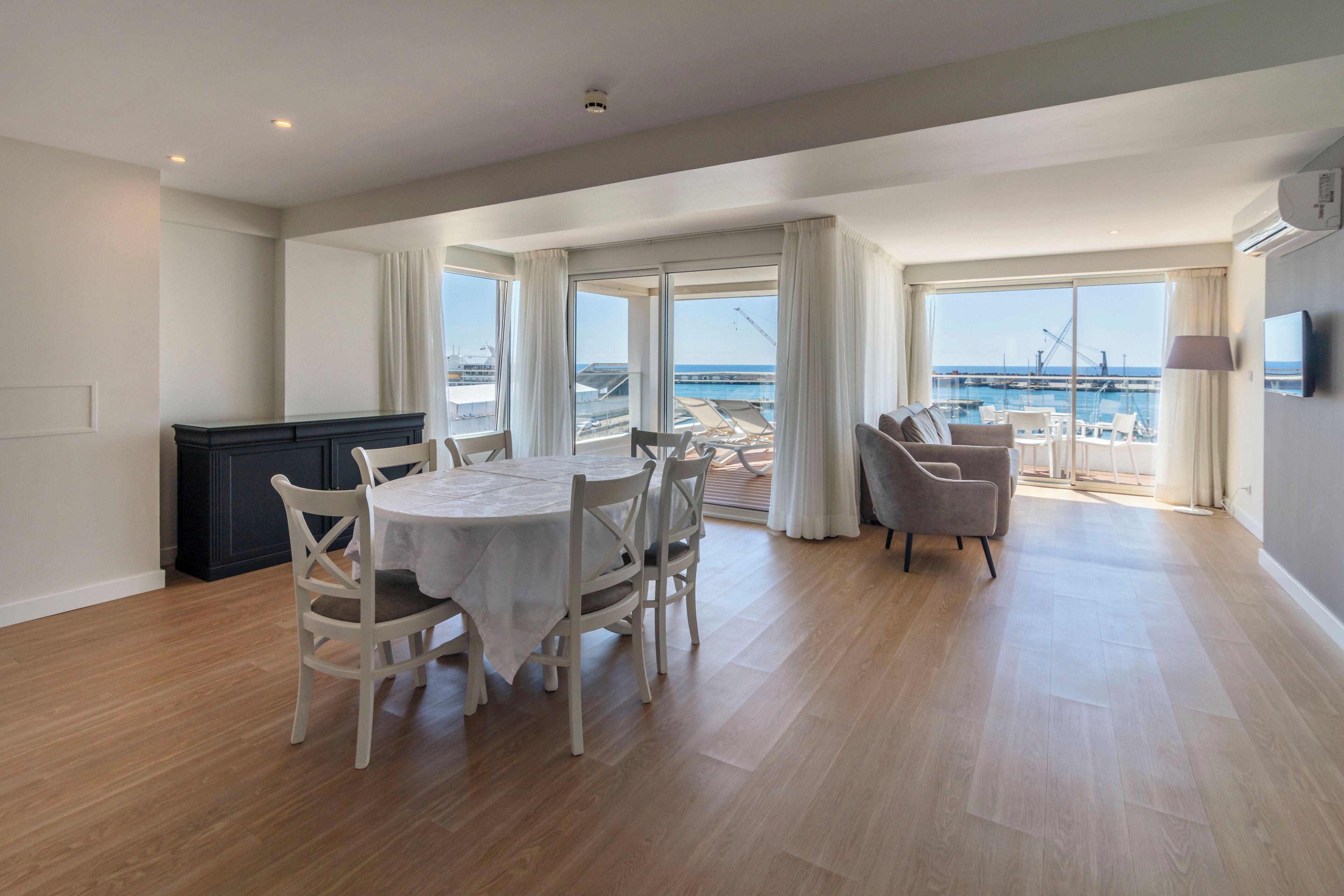 Un salón amplio con una mesa de comedor y sillas, ventanas grandes con cortinas y vistas al mar.