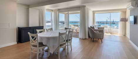 A spacious living area with a dining table and chairs, large windows with curtains, and a view of the sea.