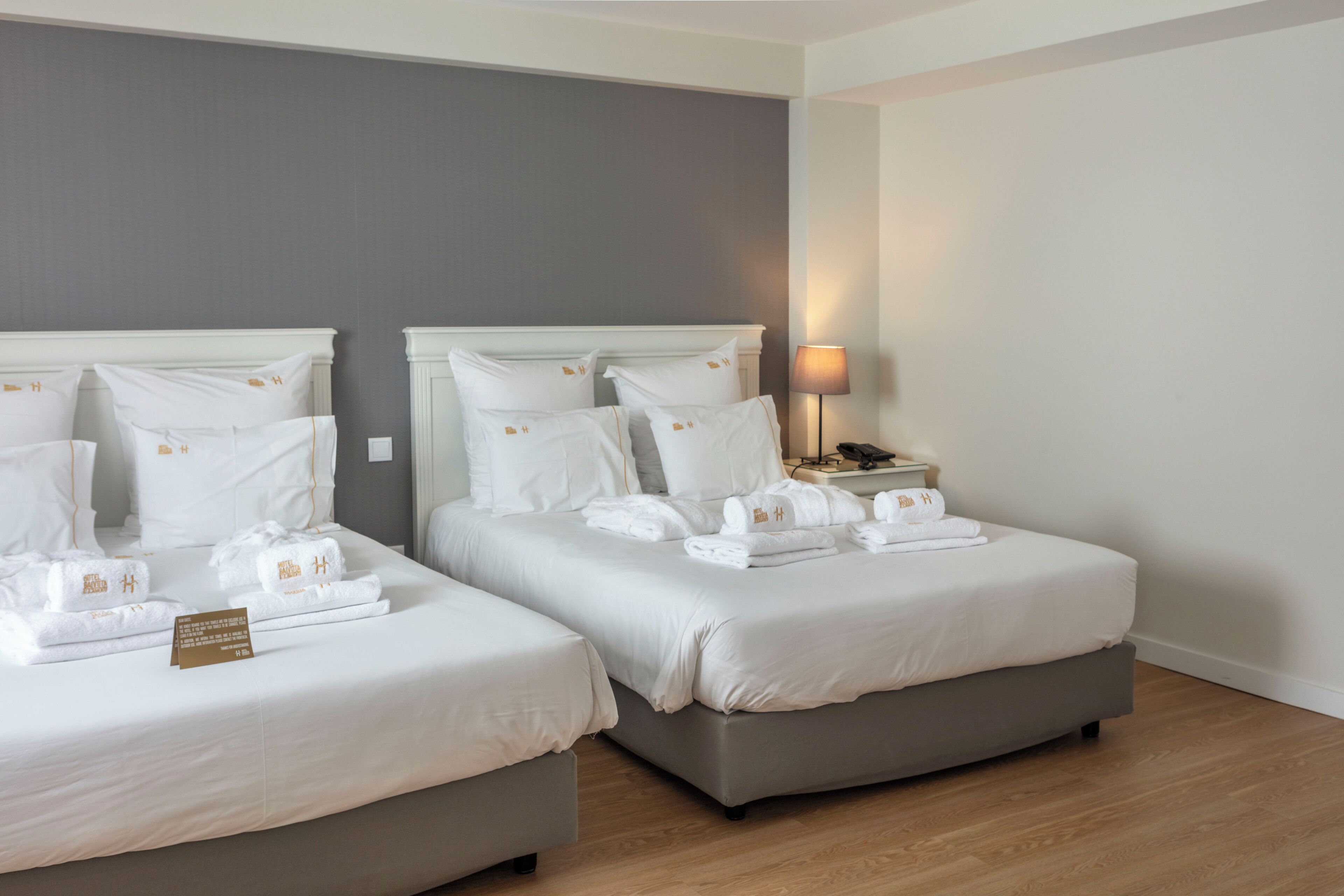 Una habitación de hotel con dos camas, cada una con sábanas y almohadas blancas, una mesita de noche con lámpara, y una tarjeta pequeña sobre la cama de la izquierda.