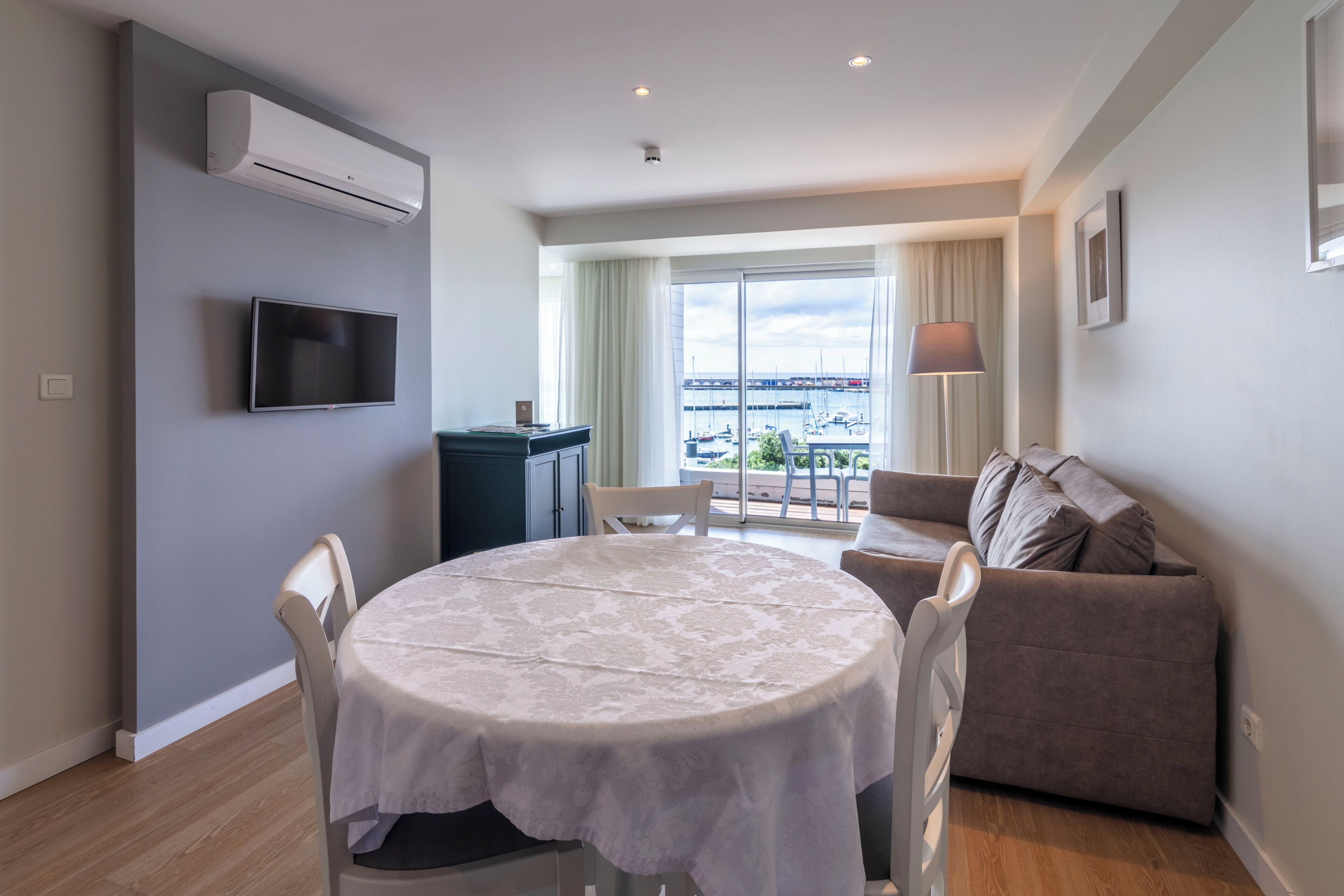 A modern living room with a round table, a flat-screen TV, a sofa, and a view of the marina through large windows.