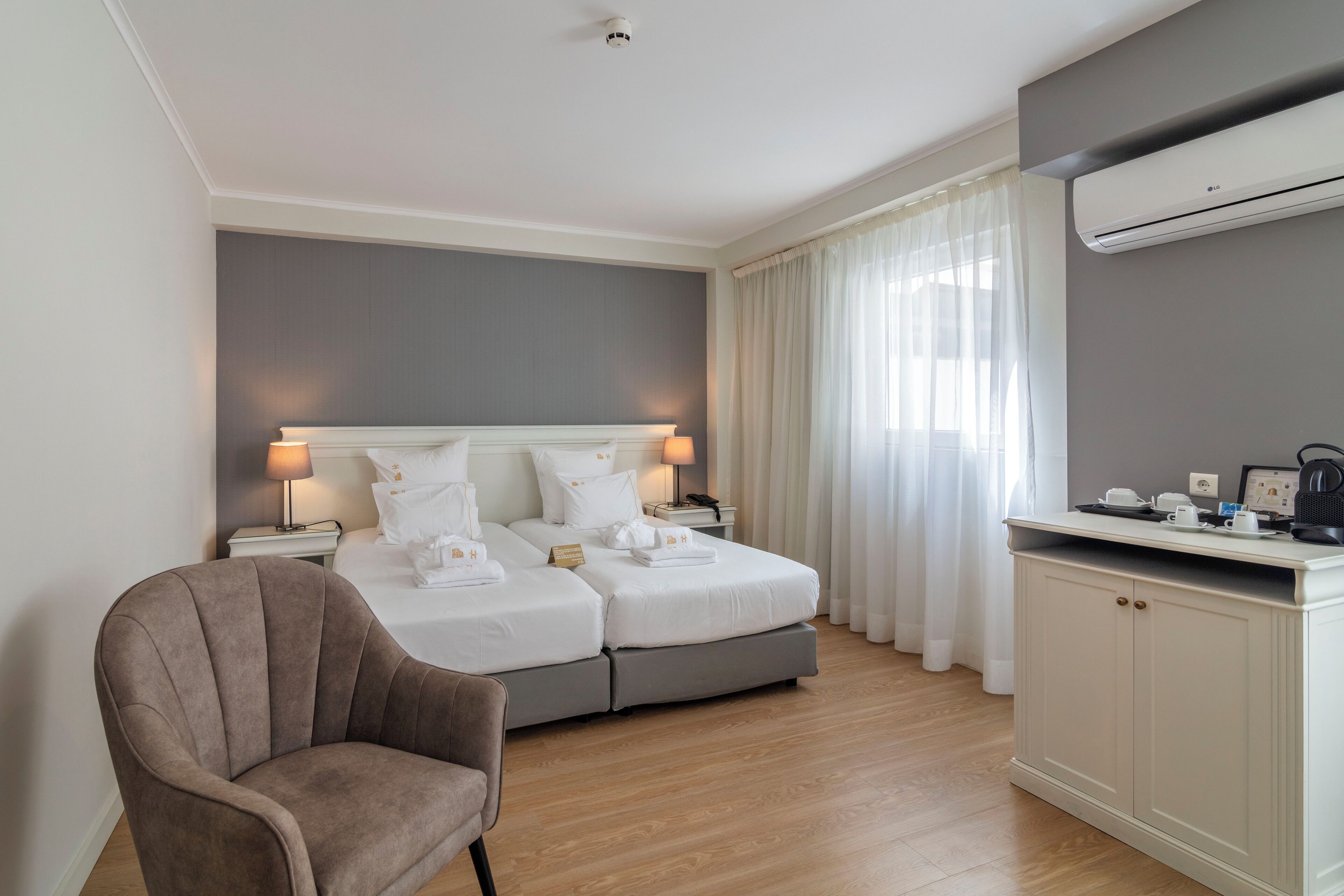 A hotel room with a bed, two bedside tables, a chair, a desk with a coffee machine, and an air conditioning unit.