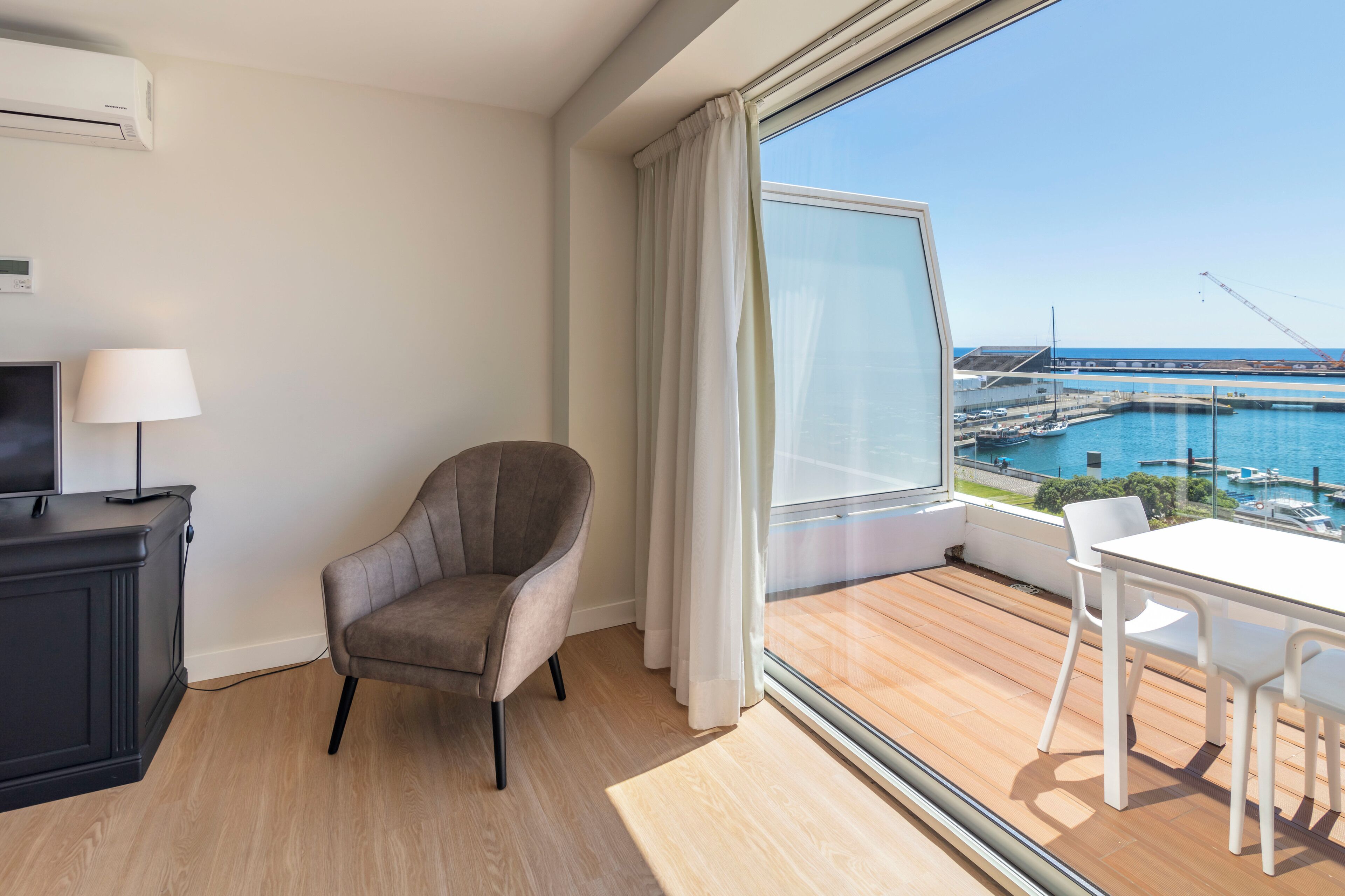A modern room with a view of a marina, a grey armchair, a white table with chairs, and a black cabinet with a lamp.