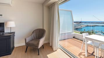 A modern room with a view of a marina, a grey armchair, a white table with chairs, and a black cabinet with a lamp.