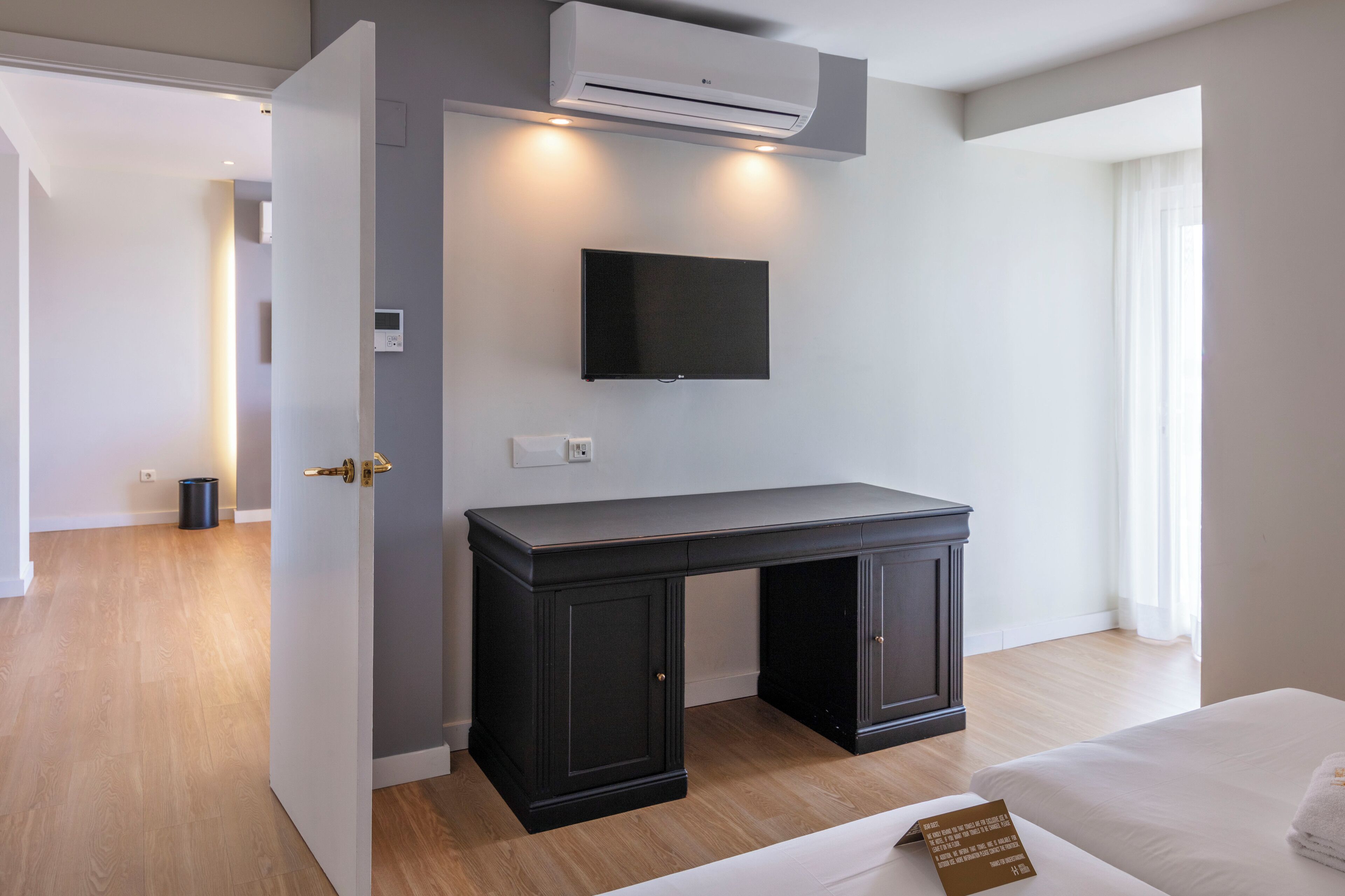 A modern hotel room with a wooden floor, a bed with a white duvet, a dark wooden desk, a mounted TV, and an air conditioning unit.