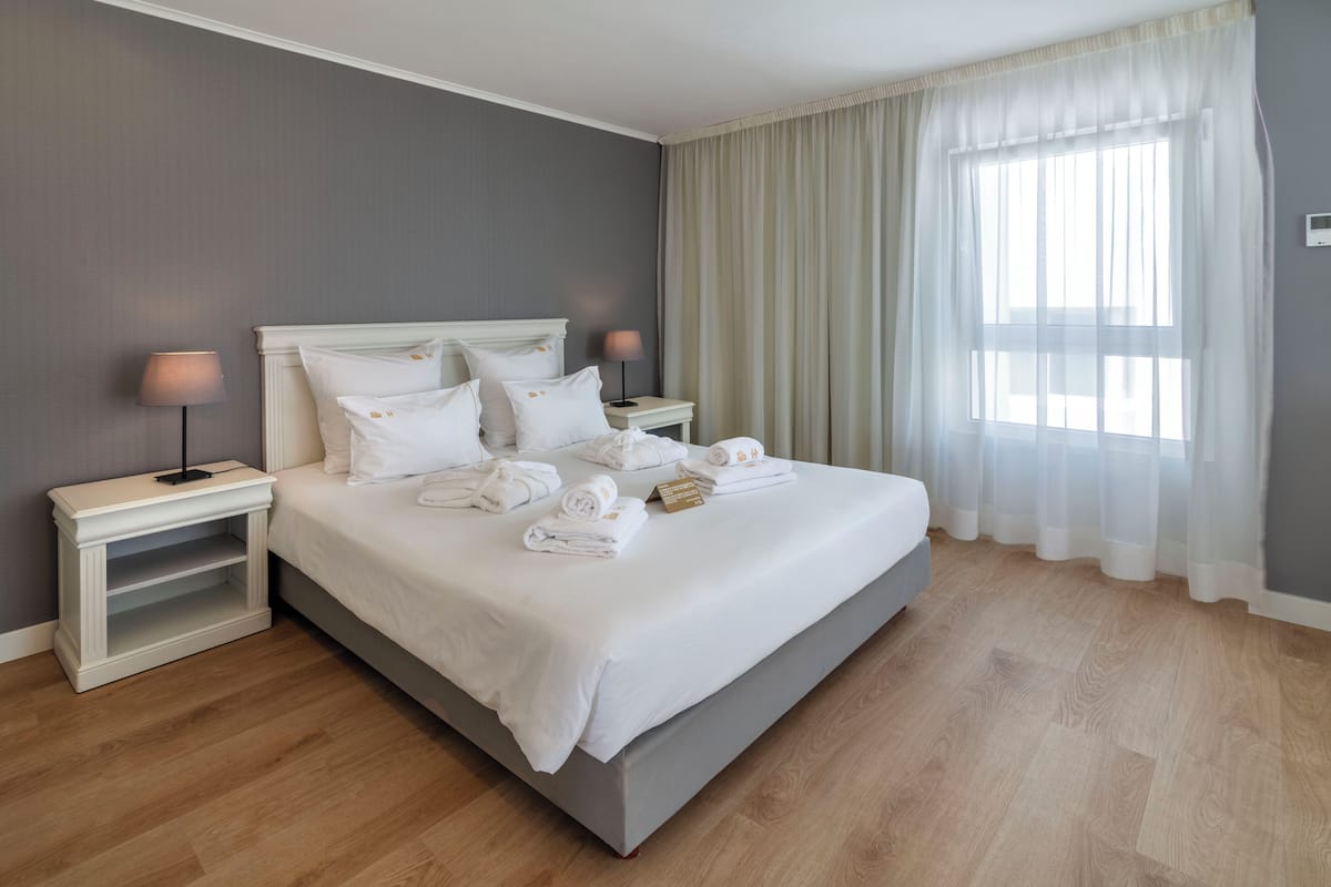 A neatly made bed with white linens and towels, a bedside table with a lamp, and a large window with sheer curtains.