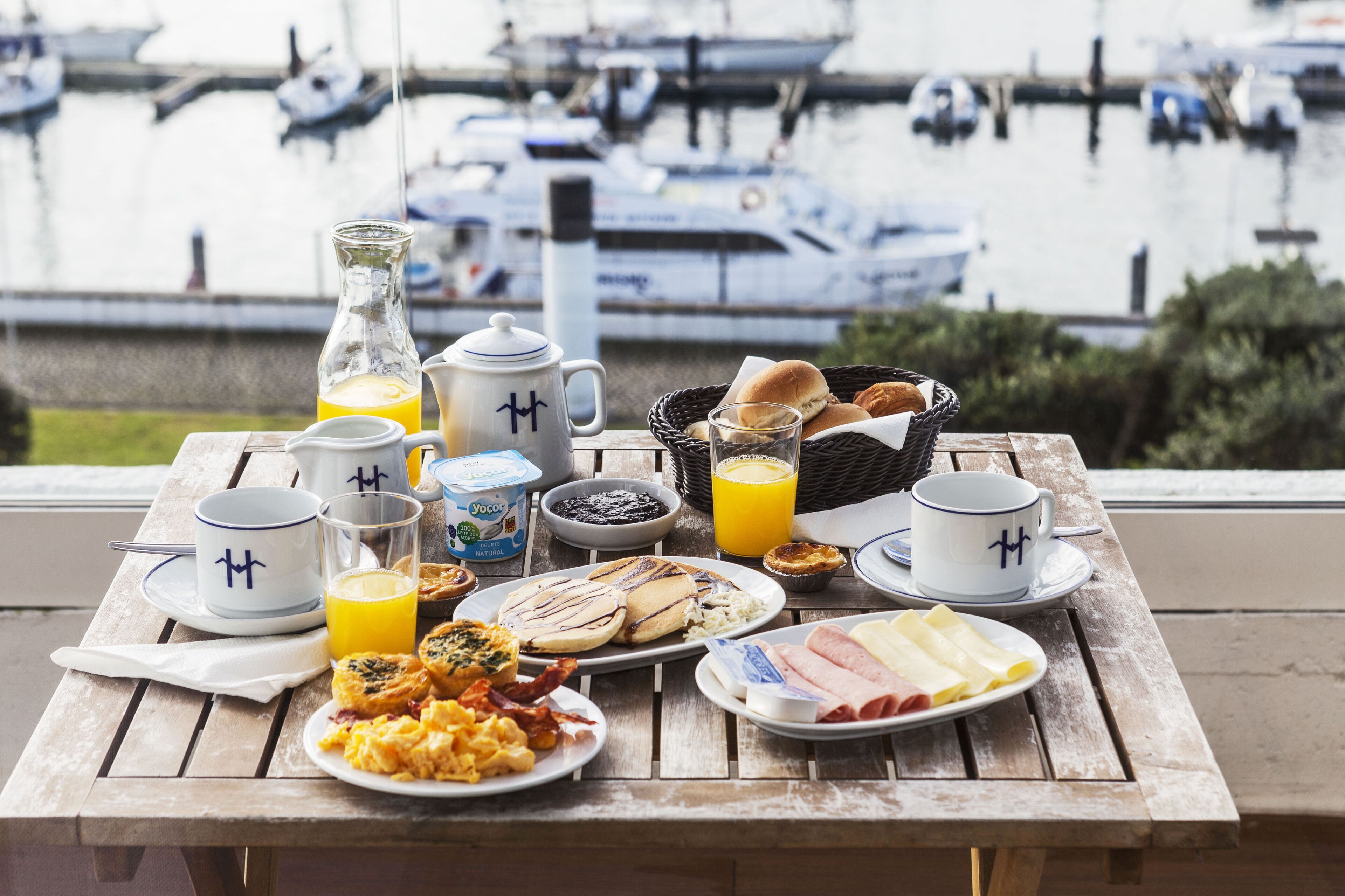 A breakfast table with various food items and drinks, overlooking a marina with boats.