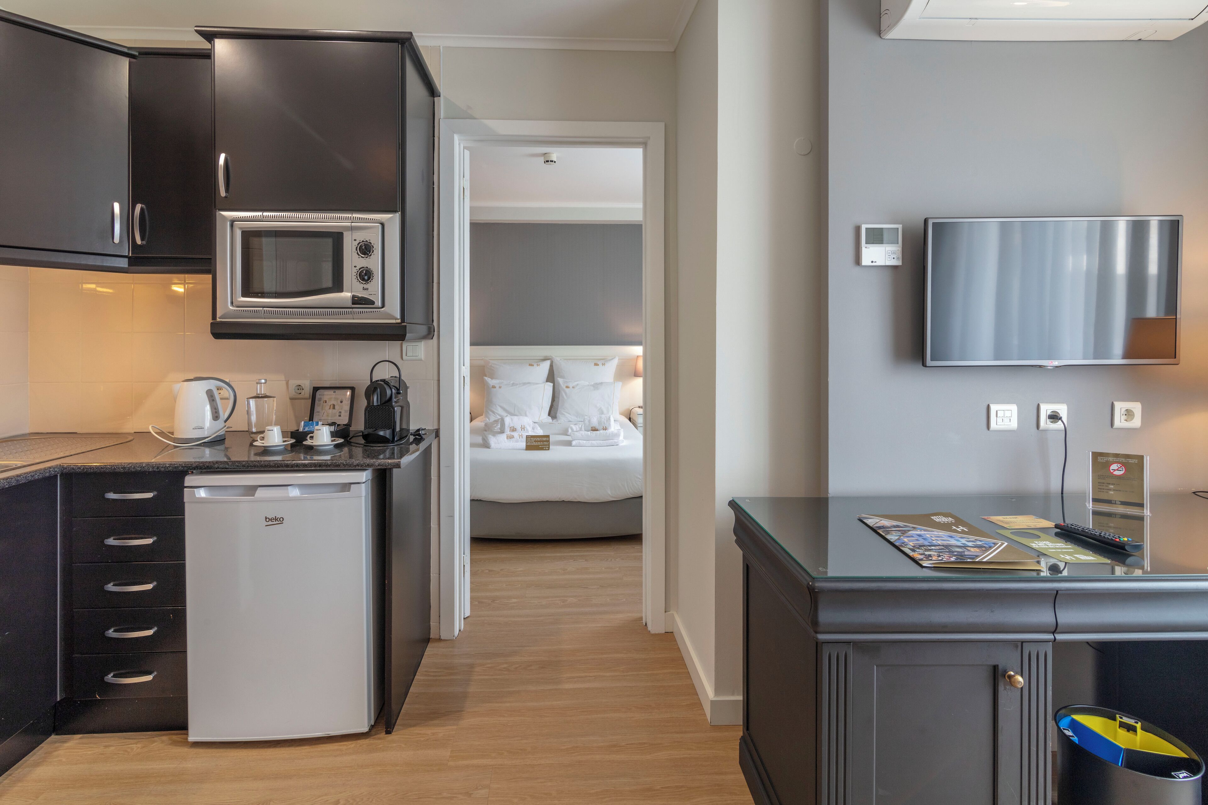 Een compacte kitchenette met een magnetron, koffieapparaat en een kleine koelkast. Door een geopende deur is een bed met wit beddengoed zichtbaar, en aan de muur hangt een flatscreen-tv.