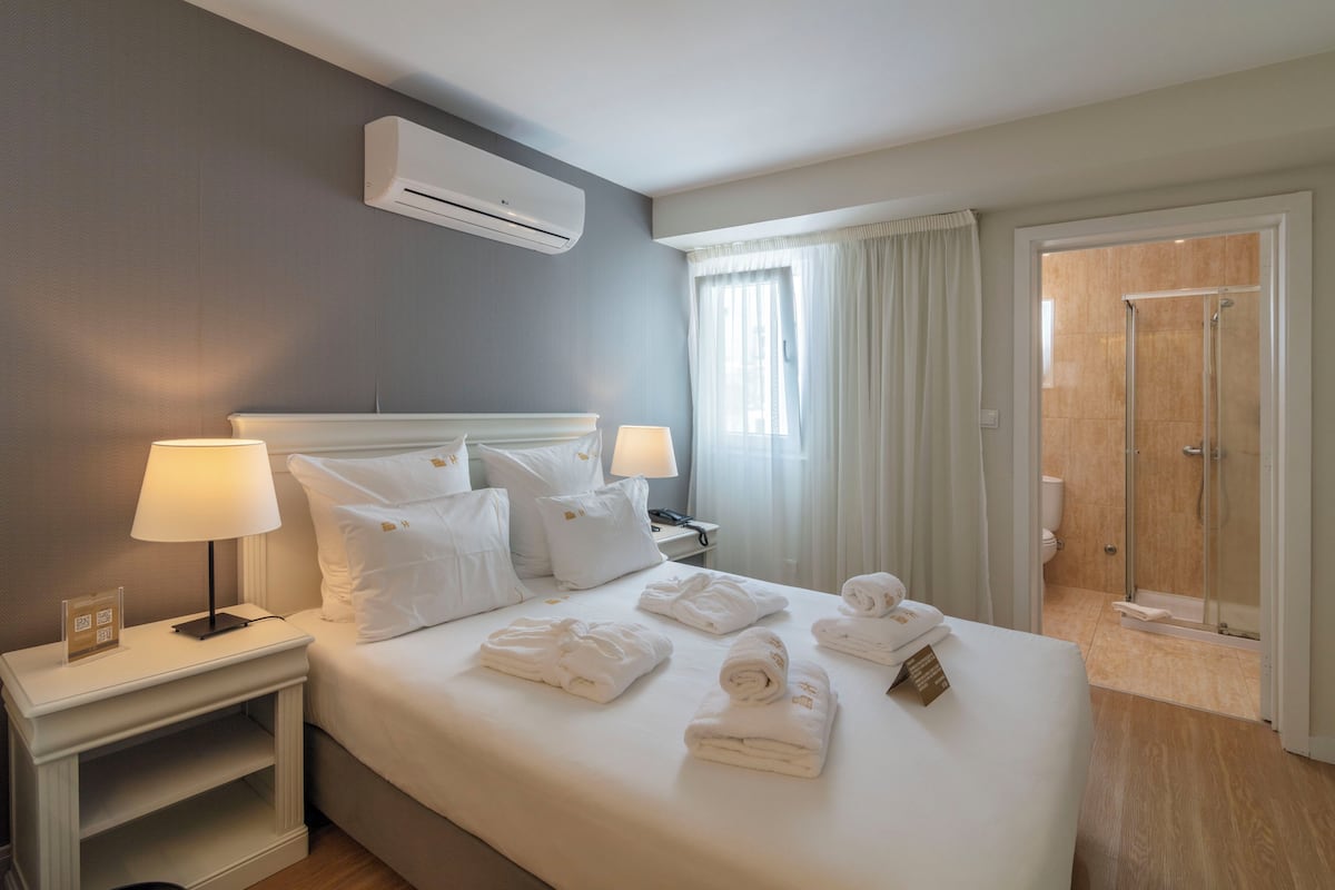 A hotel room with a bed, two bedside tables with lamps, a window with curtains, and a shower area.