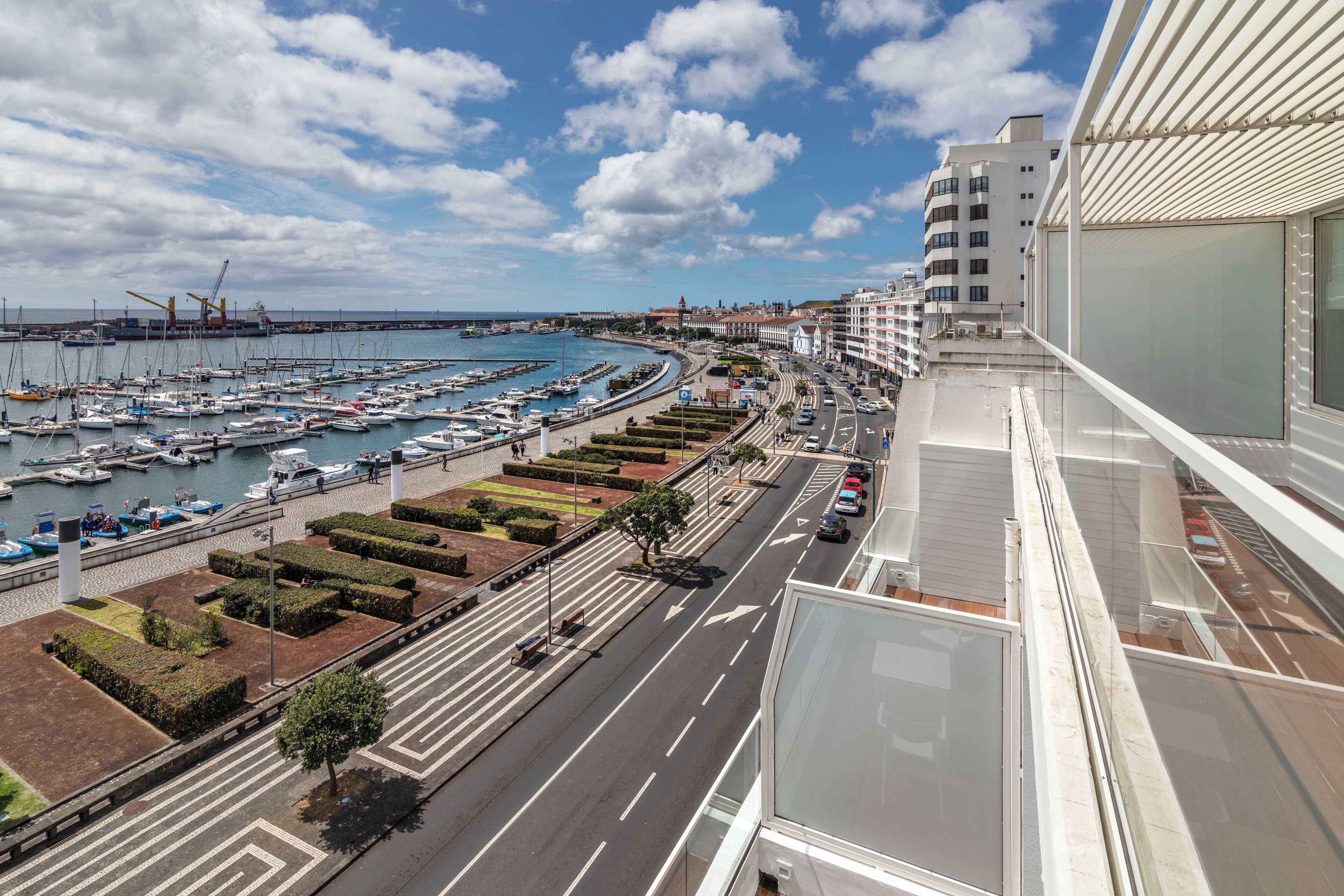 Un port esportiu amb vaixells, una carretera amb cotxes i edificis al fons.