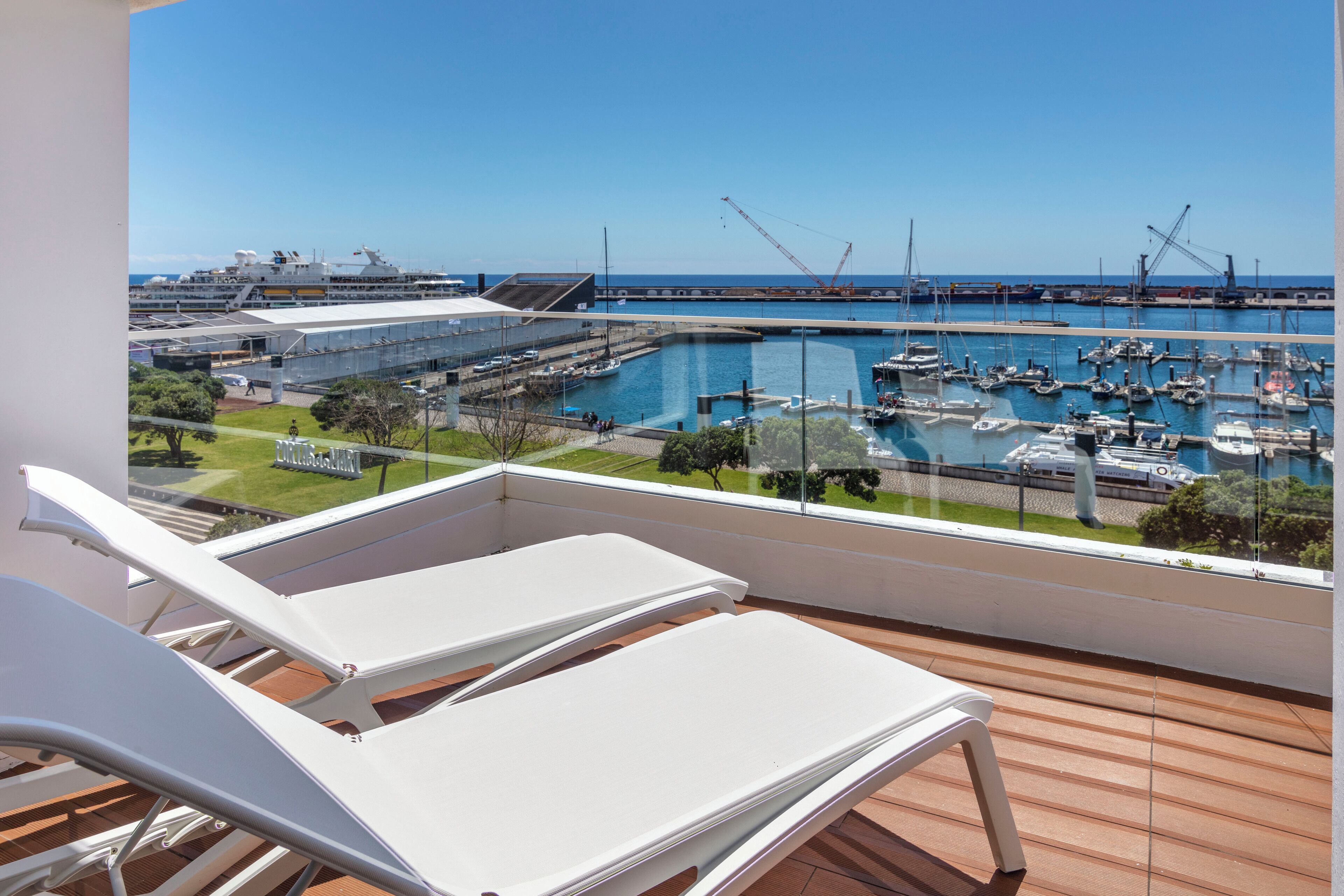 Un balcón con sillones blancos que da a un puerto deportivo con barcos y un crucero amarrado al fondo.