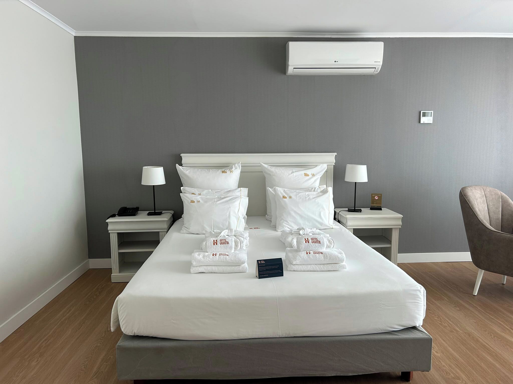 A hotel room with a bed, two bedside tables, a chair, an air conditioning unit, and a thermostat.