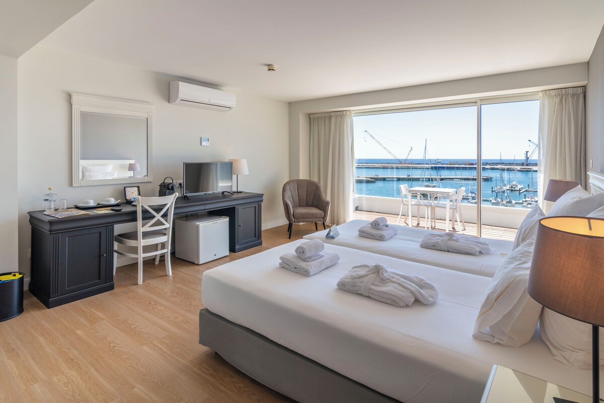 A hotel room with a large bed, a desk, a chair, and a view of the marina.
