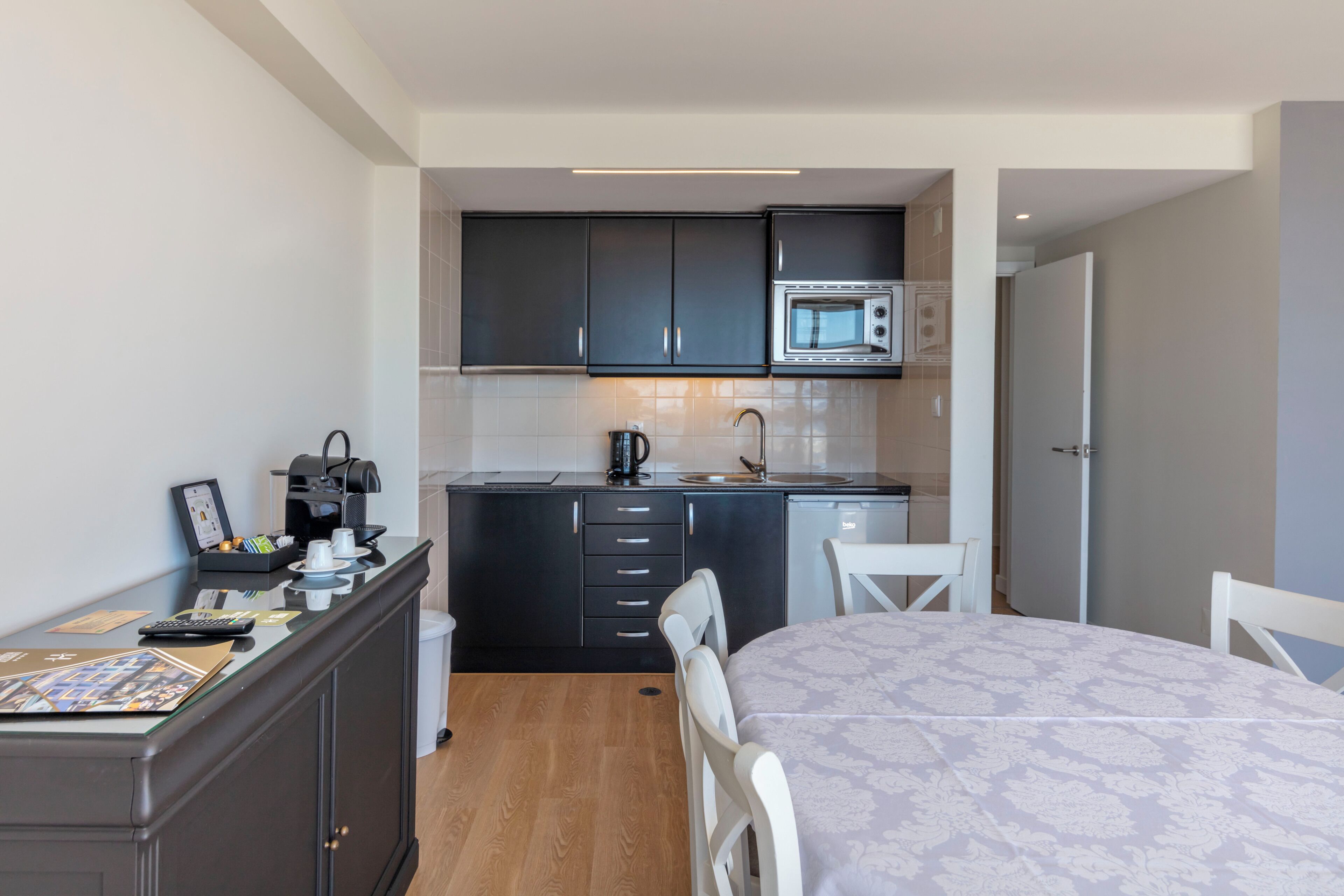 Een compacte keuken met donkere kastjes, een magnetron en een koffieapparaat. Er is een eettafel met stoelen en een klein aanrecht met een koffiehoek.