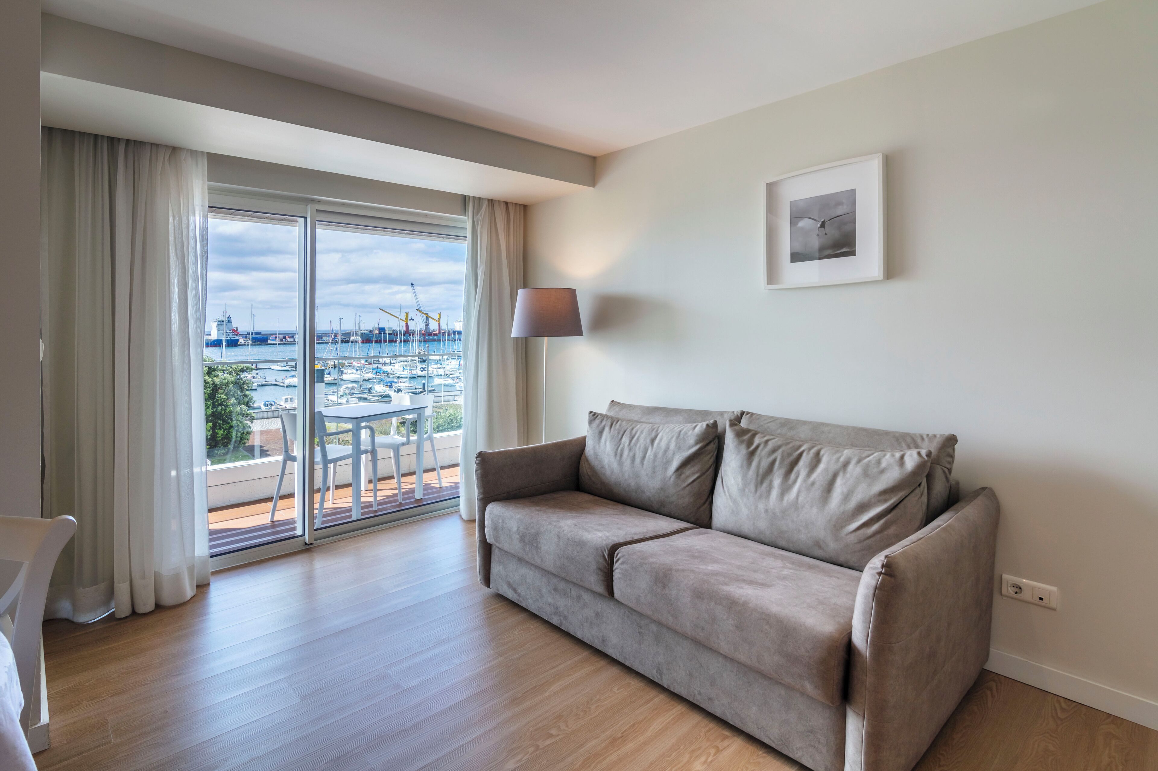 A modern living room with a grey sofa, a table, and a chair. A large window offers a view of a marina and boats.