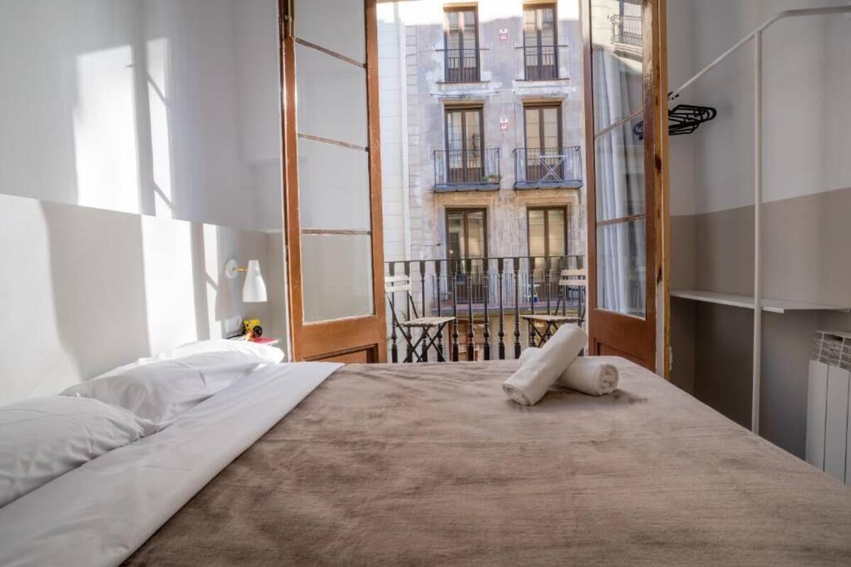 A hotel room with a bed, a nightstand, a window with a view of a building, and a balcony with a railing.