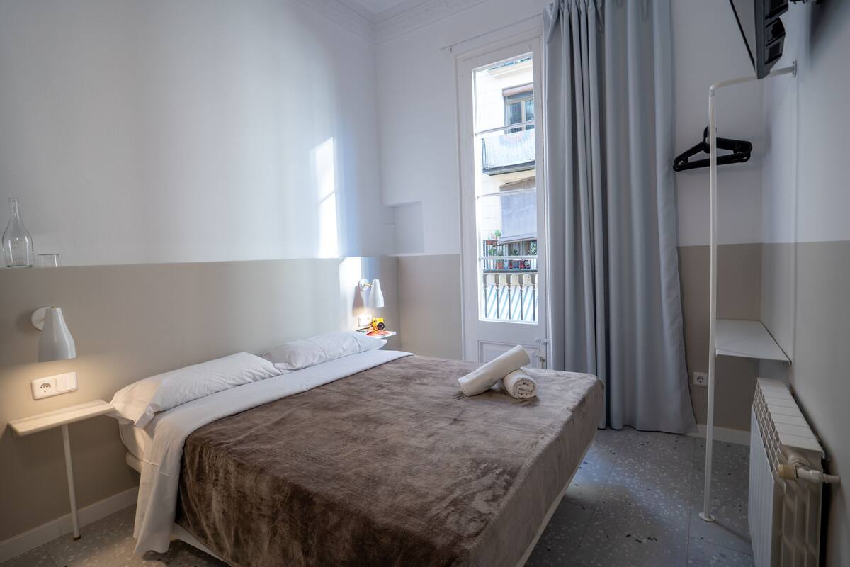 A hotel room with a bed, a bedside table with a lamp, a window with curtains, and a radiator.