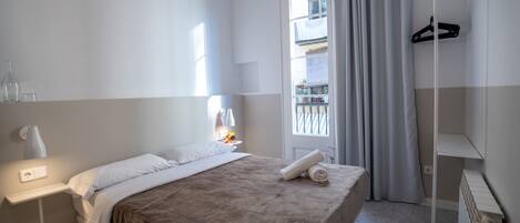 A hotel room with a bed, a bedside table with a lamp, a window with curtains, and a radiator.
