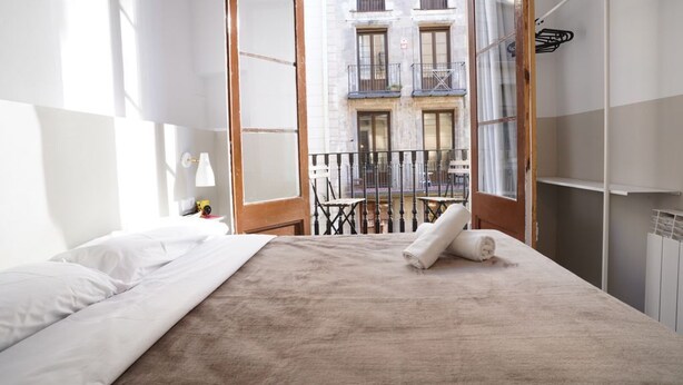 A hotel room with a bed, a nightstand, a lamp, and a view of a building with balconies. - Hostal Hera (Barcelona)