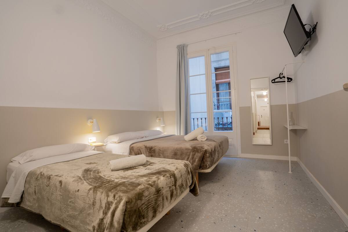 A hotel room with two beds, a TV mounted on the wall, a window with a view of a balcony, and a mirror on the wall.