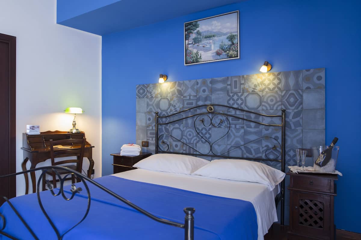 A hotel room with a bed, a desk, a chair, a nightstand, and a wall-mounted lamp.