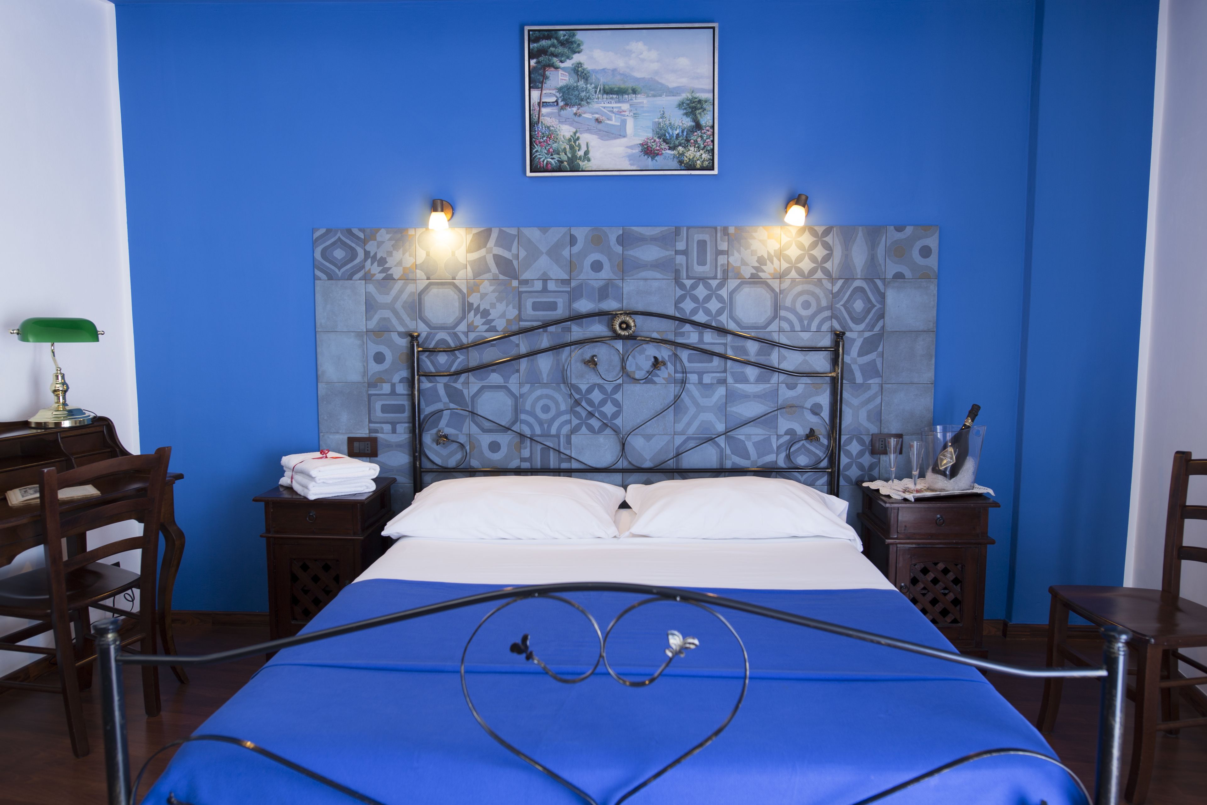 Ein Hotelzimmer mit blauer Wand, einem Bett mit Metallkopfteil, zwei Nachttischen mit Lampen, einem Bild an der Wand und Blick auf eine Landschaft.