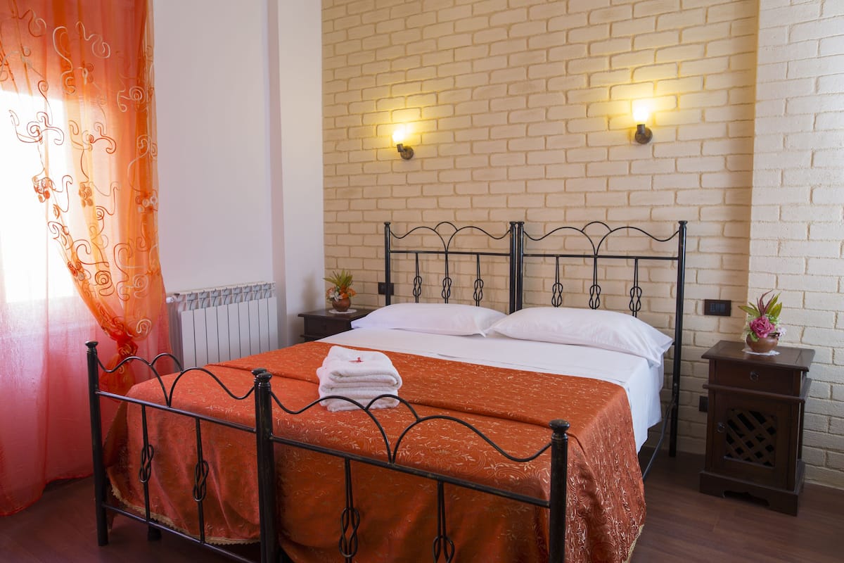 A double bed with an orange bedspread, a white pillow, and a folded white towel on the headboard. There is a nightstand with a small plant and a lamp. The room has a brick wall and a window with orange curtains.