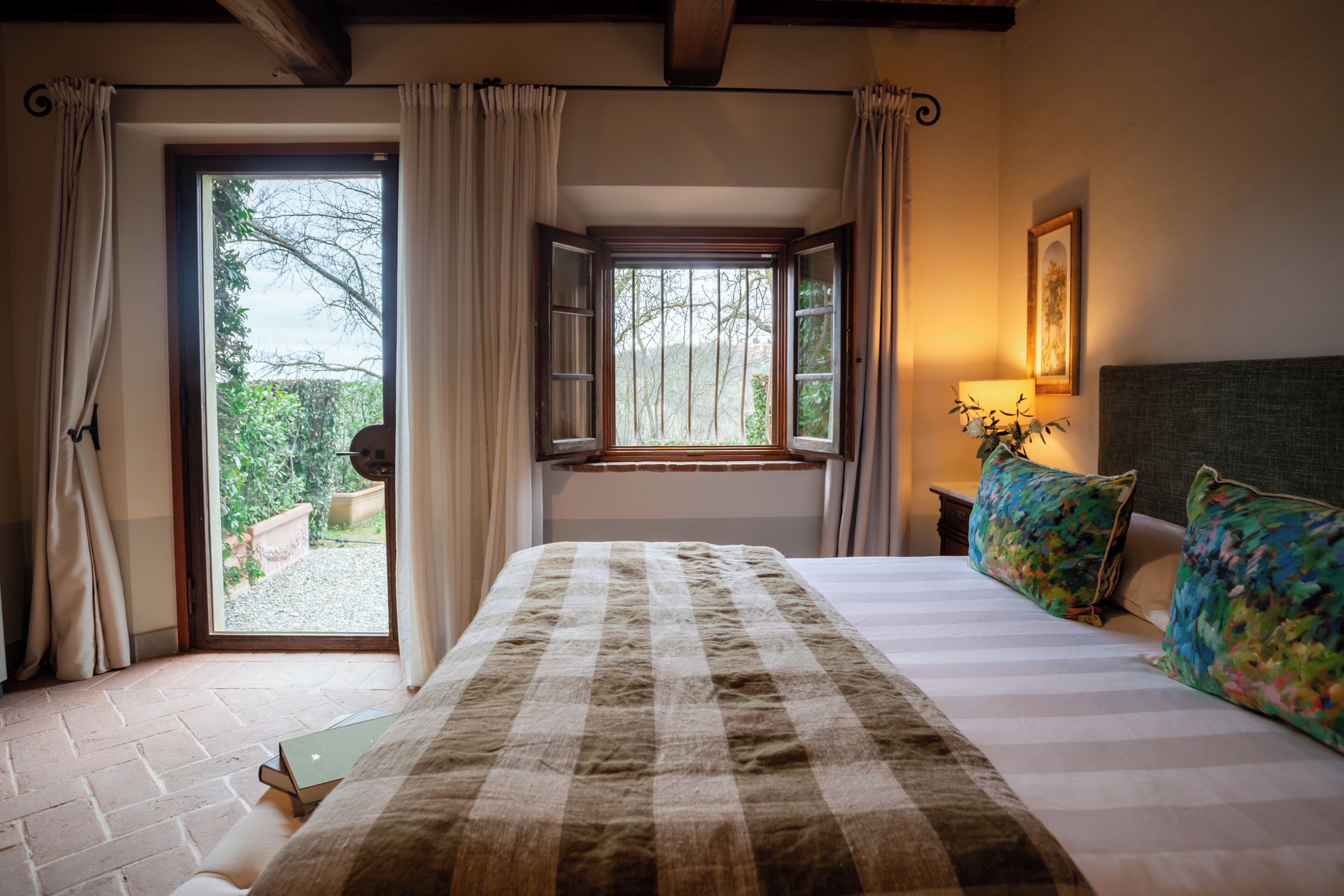 Ein Schlafzimmer mit einem großen Bett, einem Fenster mit Vorhängen, einem Nachttisch mit Lampe und einem gerahmten Bild an der Wand.