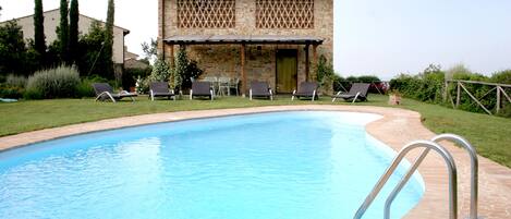 Una piscina con plataforma de piedra, rodeada de sillas de descanso y un edificio de piedra con techo de tejas.