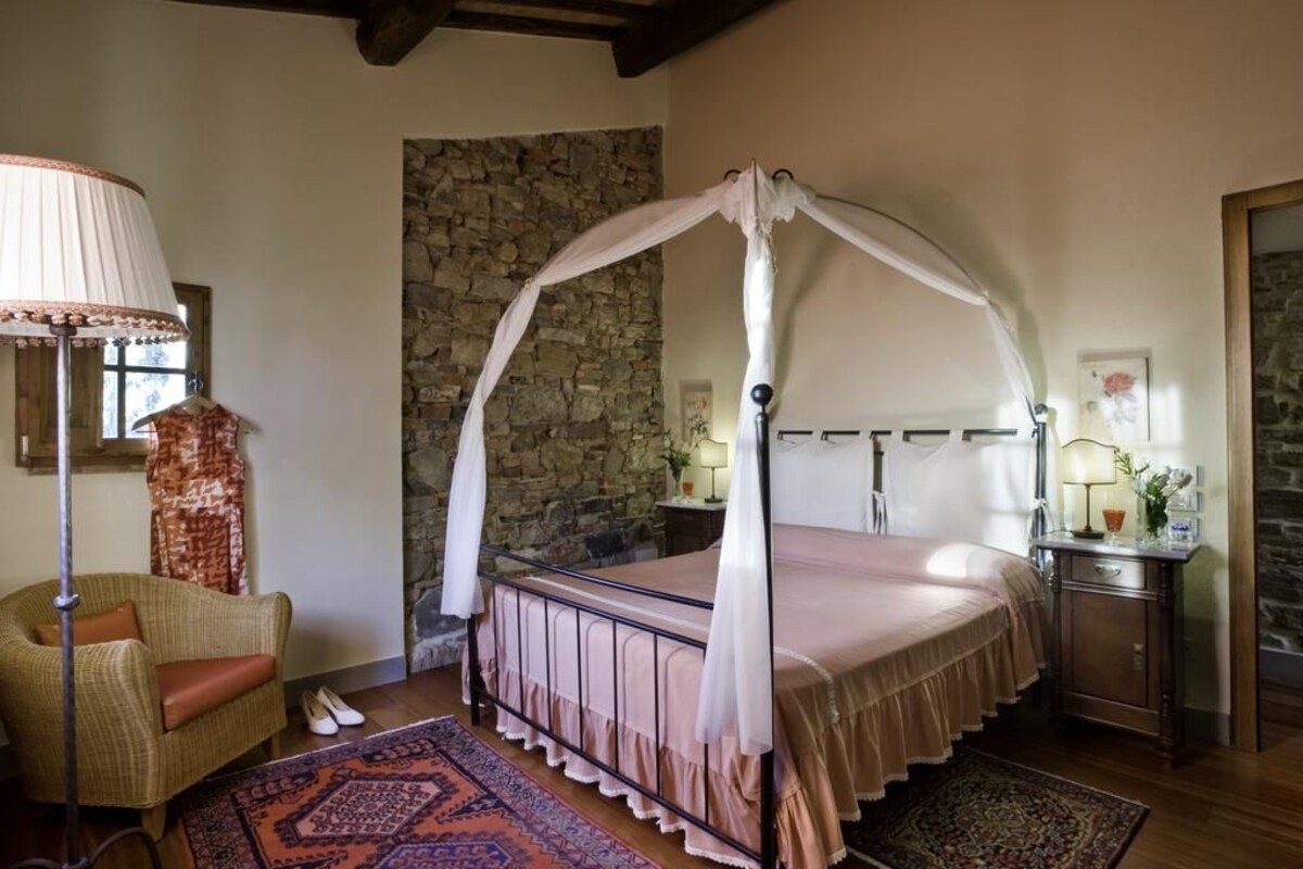 A bedroom with a four-poster canopy bed, a stone wall, a wicker chair, and a nightstand with a lamp.