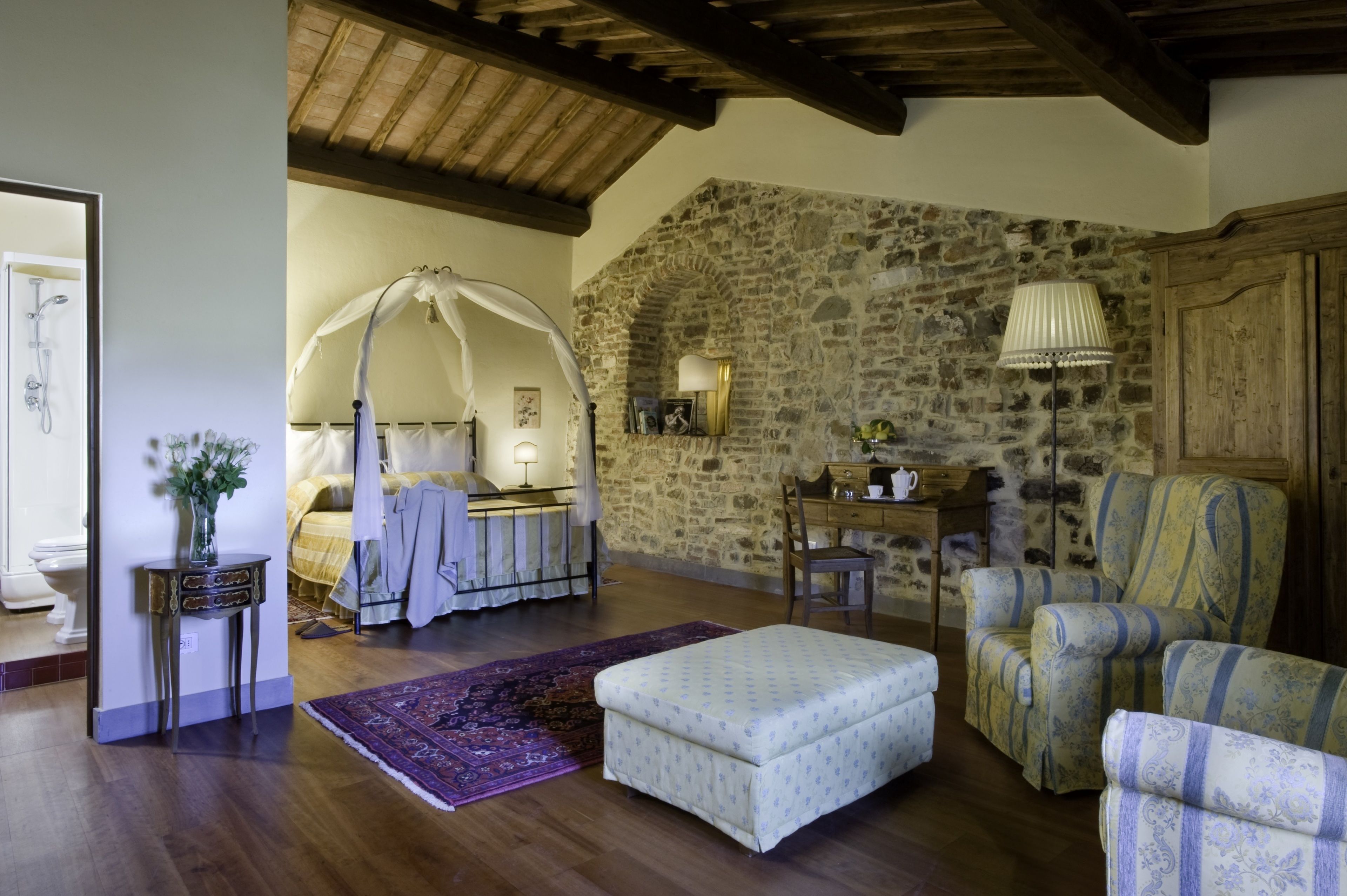 A bedroom with a stone wall, a canopy bed, a bedside table with a lamp, and a chair.
