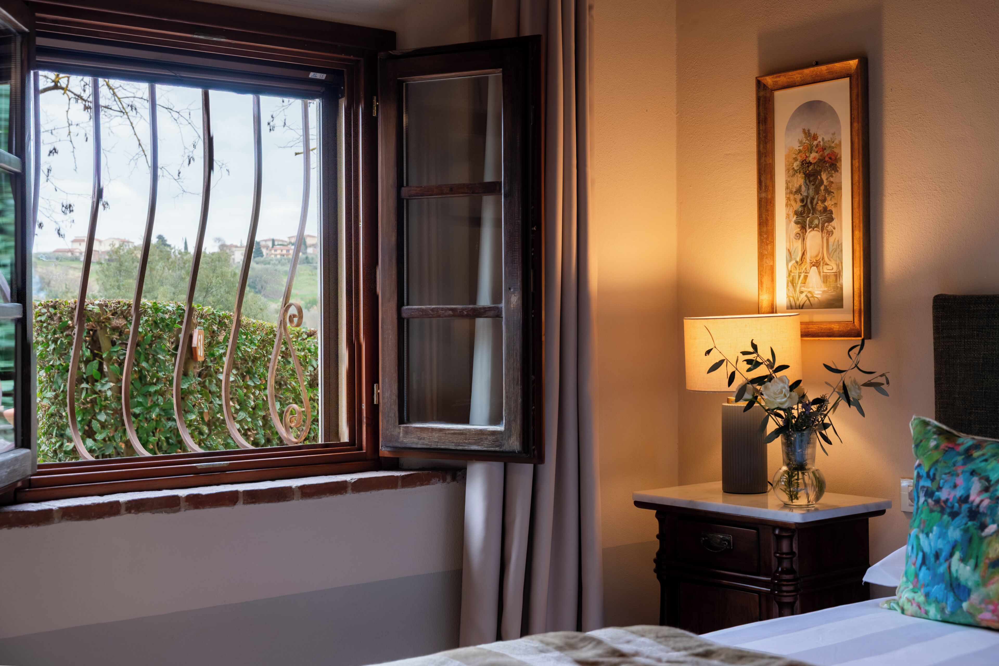 A hotel room with a bed, a bedside table with a lamp, a window with a view of greenery, and a framed picture on the wall.