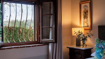 Habitación de hotel con cama, mesita de noche con lámpara, ventana con vista a follaje y un cuadro en la pared.