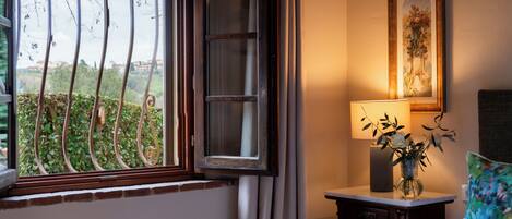 A hotel room with a bed, a bedside table with a lamp, a window with a view of greenery, and a framed picture on the wall.