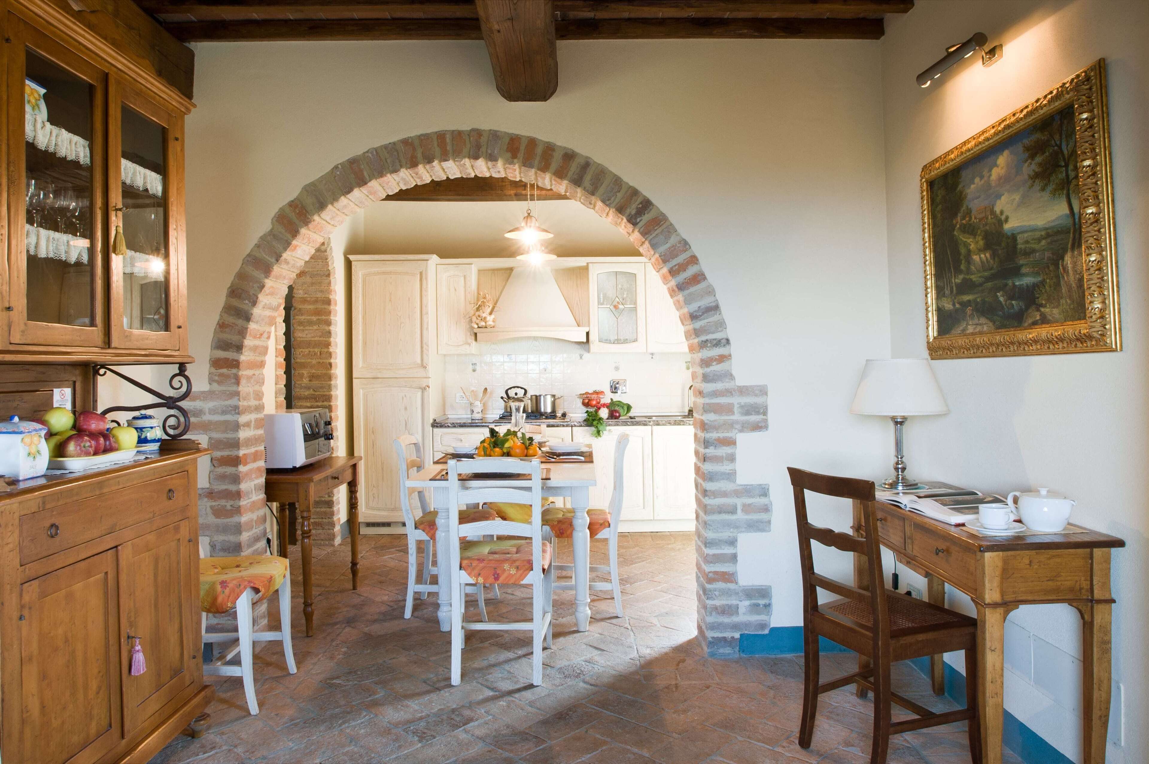 A rustic kitchen with an archway leading to a dining area, featuring a wooden table, chairs, and a painting on the wall.