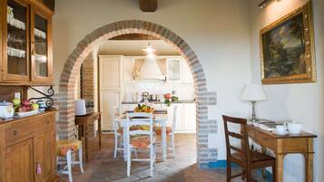 A rustic kitchen with an archway leading to a dining area, featuring a wooden table, chairs, and a painting on the wall.