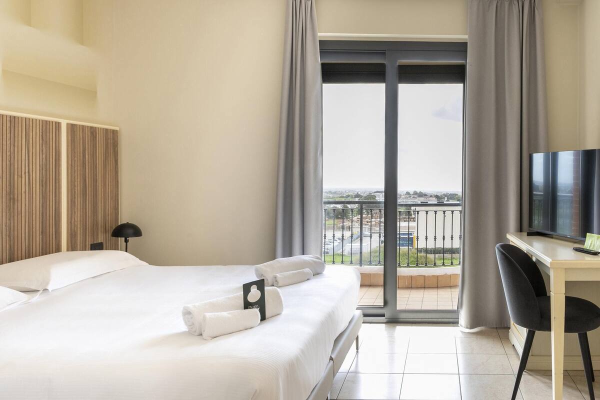 A hotel room with a bed, a desk, a television, and a balcony with a view of the city.