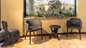 A seating area with two chairs and a small table, a decorative sculpture, and a window with a view of a garden and buildings. - B&B Hotel Pomezia Cortese (Pomezia)