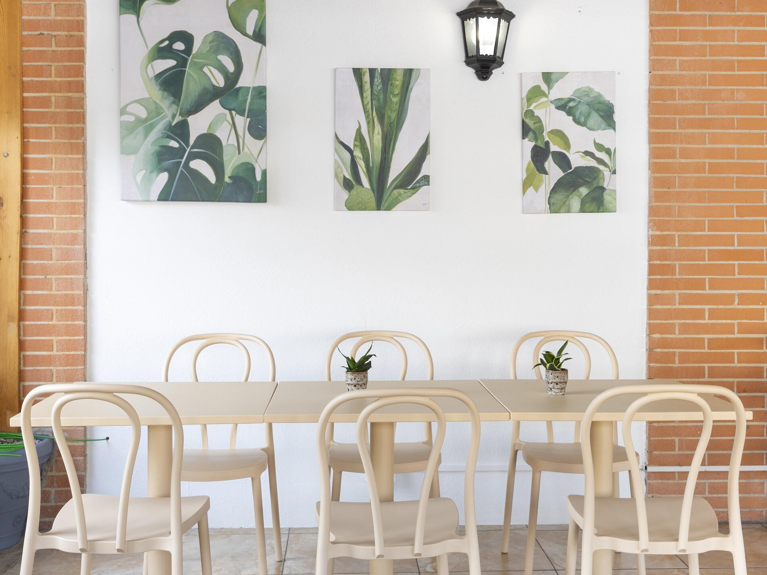 A dining area with a long table and six chairs, three potted plants on the table, and three framed botanical prints on the wall.