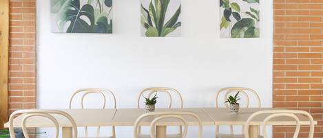 A dining area with a long table and six chairs, three potted plants on the table, and three framed botanical prints on the wall.