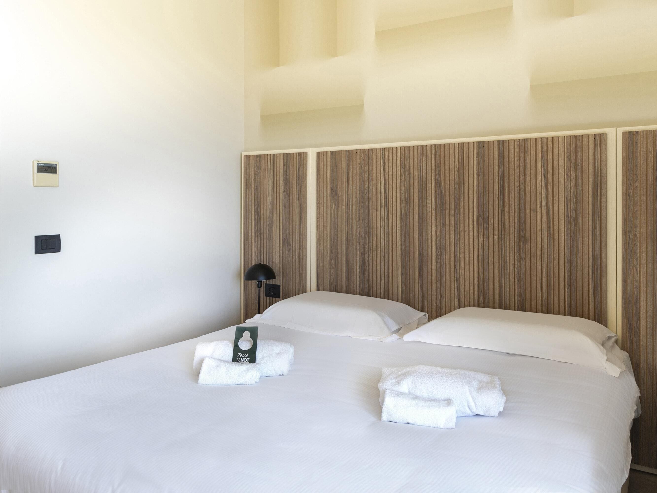 Quarto de hotel com uma cama grande, duas almofadas, mesa de cabeceira com abajur e painel de controle fixado na parede.