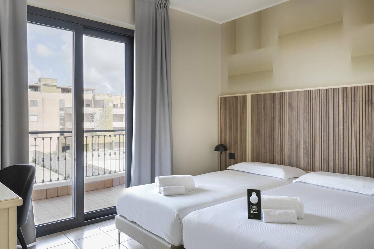 A hotel room with two beds, a desk, a chair, a balcony with a view of buildings, and a sign on the bed.