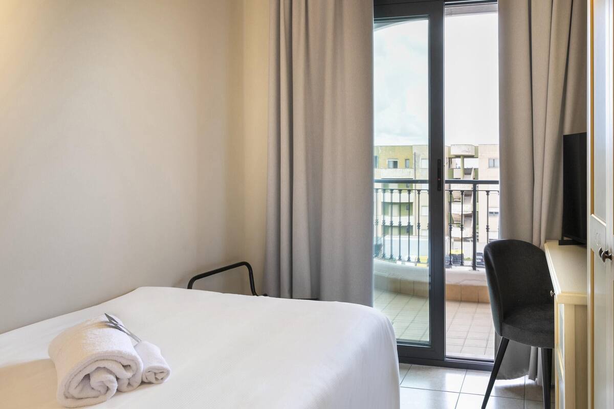 A hotel room with a bed, a chair, a desk, and a balcony with a view of buildings.