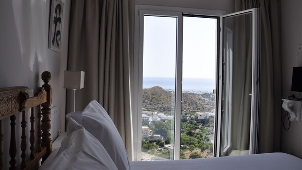 A hotel room with a bed, a wooden headboard, a bedside lamp, a TV, and a large window with curtains. - Hotel Mamabels (Mojacar)