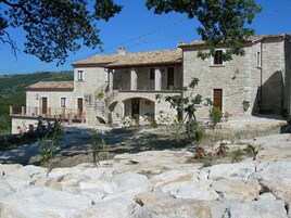 Un edificio de piedra con techo de tejas, una escalera que conduce a un balcón y un patio empedrado con rocas grandes.